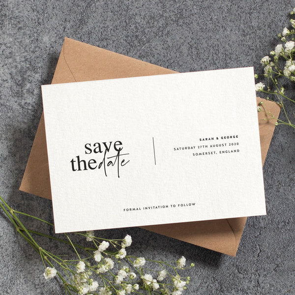 A white 'save the date' card on a brown box with babys breath laying beside the box
