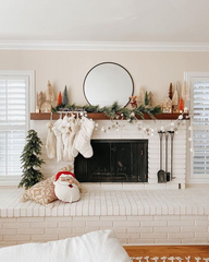 Boho Christmas Mantel. Cream coloured living room with an off-white brick fireplace and mantel. There are white stockings hanging on the left side, a garland of white balls and another garland of silver/gold snowflakes hanging across the mantel. On top is a small garland of pine, neutral coloured festive house sculptures, and neutral brush bottle trees with a pop of orange trees. There is a circular mirror with a black rim in the centre of the mantel.