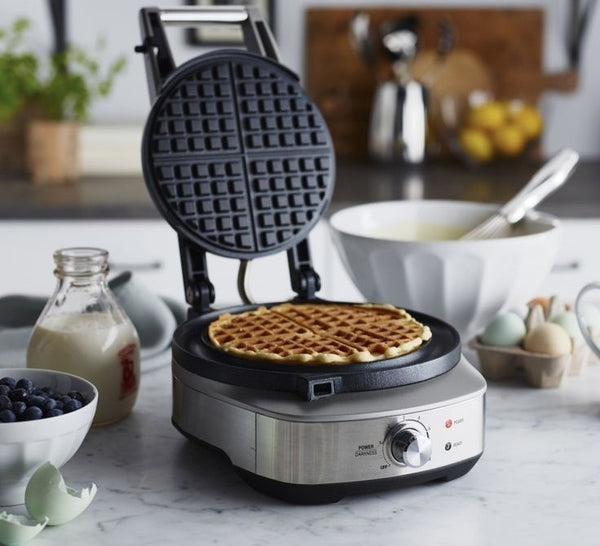 A waffle maker, with a waffle inside, is on a marble table. Baking ingredients surround it. Modern home with a blurred background.