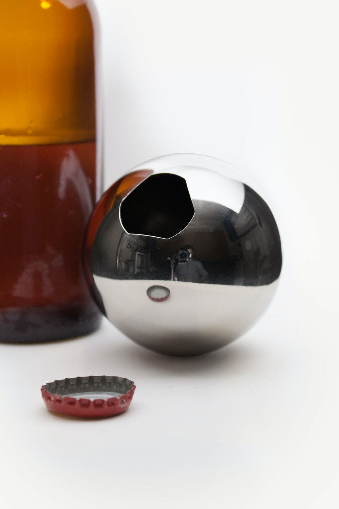 A stainless steel Convex bottle opener sitting on a white background next to a brown bottle and red bottle cap.