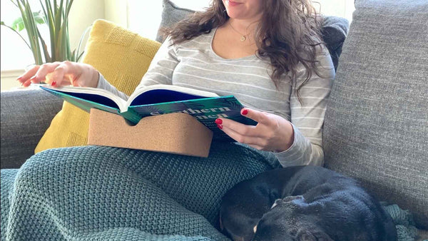 Revision Cork booklift holds book while Woman pages through large book on grey couch with green throw blanket.