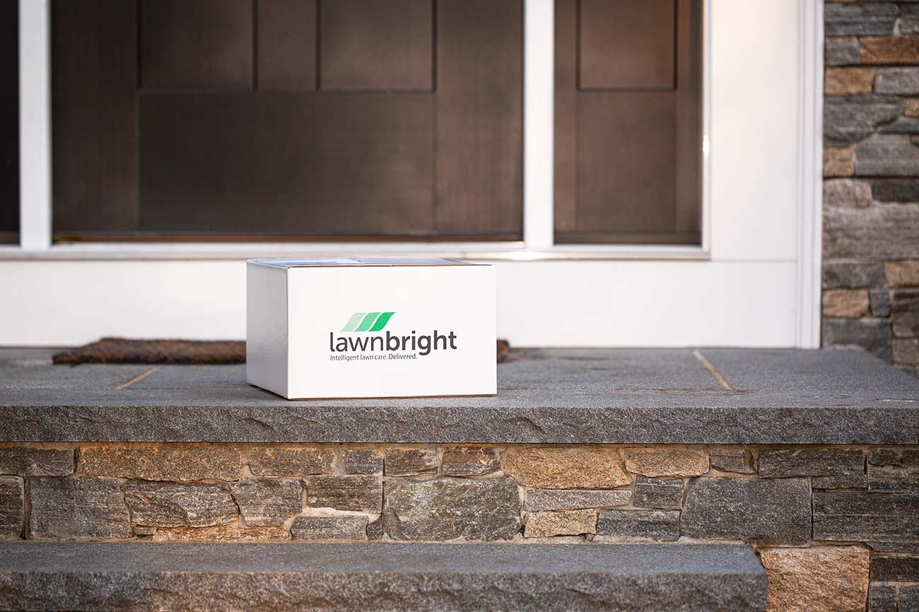 A Lawnbright branded box sits on a stone porch in front of a house with a large window.