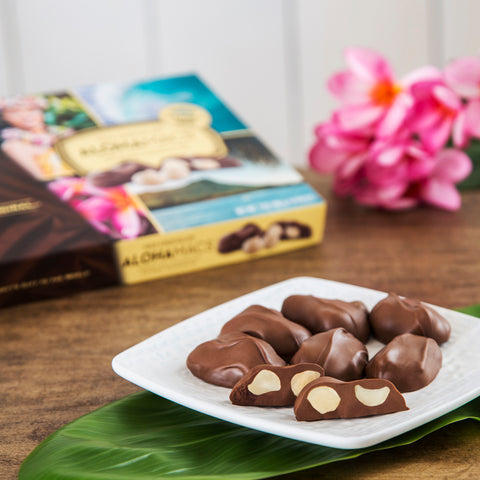 Hawaiian Host AlohaMacs Milk Chocolates ona white plate with a green ti leaf with pink plumeria flowers on the side and packaging.