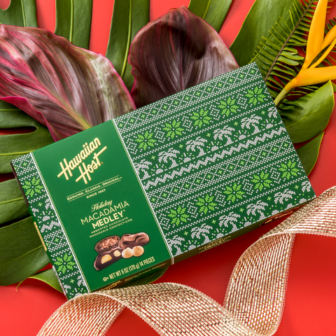 Hawaiian Host Holiday Macadamia Medley Box on red background on top of tropical flowers like monstere leaf birds of paradise and gold ribbon