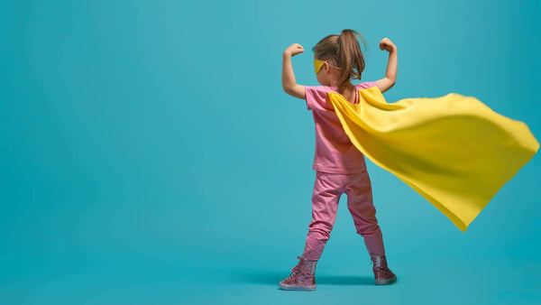 Girl wearing a yellow cape in a superhero pose against a turquoise background