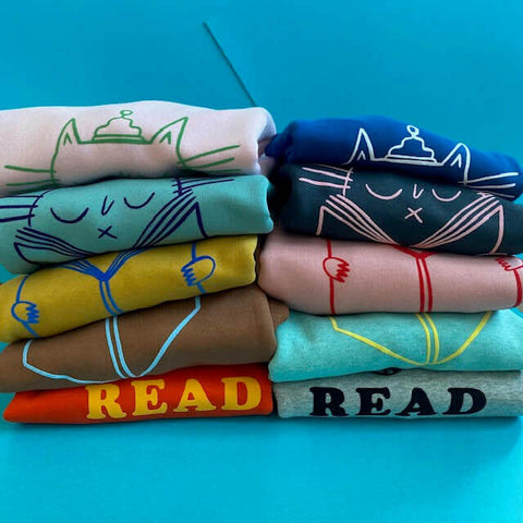 Piles of folded sweatshirts showing an illustration of a cat reading and the word READ