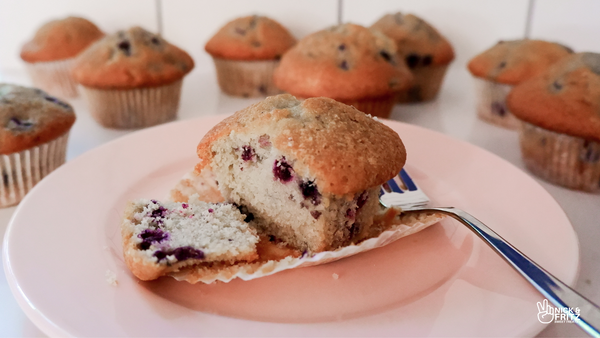 Photo from the front of a half-eaten blueberry muffin on a pink plate with more muffins in the background