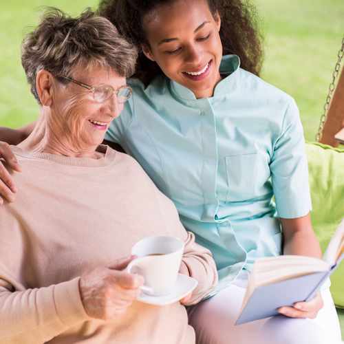 two women smiling reading a book, one is a senior woman and the other is a young caregiver