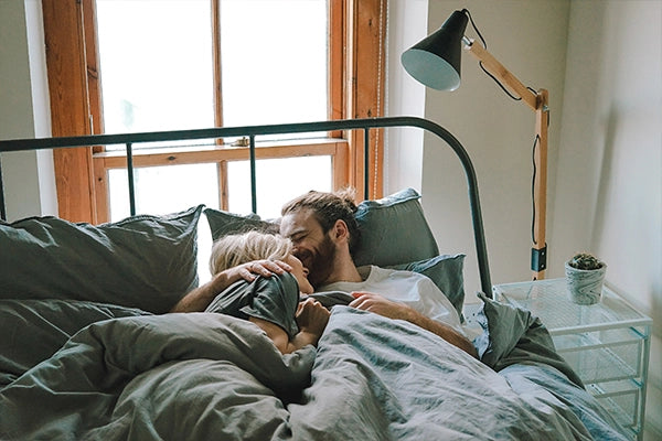 Woman with blonde hair and man with brown hair and beard cuddling in green bed sheets