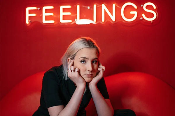 Woman with blonde hair sitting with chin in hands in front of neon sign that reads "feelings"