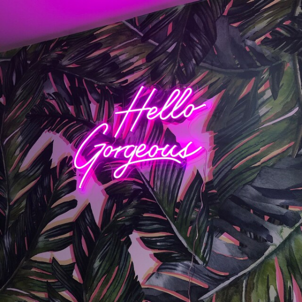 Customer's Hello Gorgeous sign in neon pink. The sign is installed in the middle of a wall. The wall is covered in a palm tree pattern wallpaper.