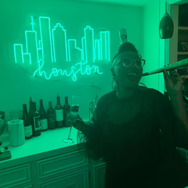 Customer's photo of their green Houston Skyline Neon. They are posing infront of it whilst holding a drink. Their neon sign installed above their home bar.