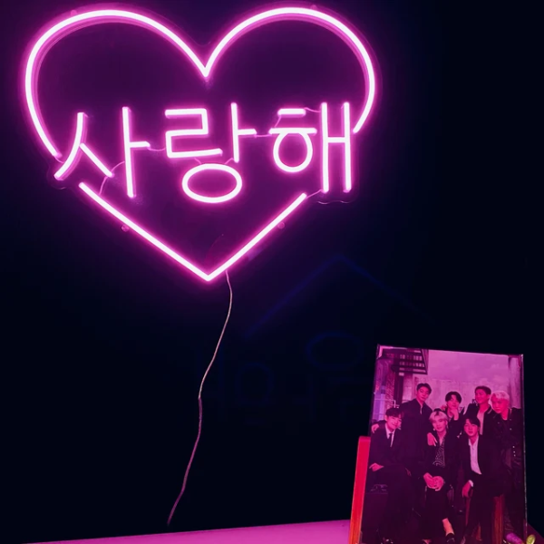 Love you in korean inside a heart neon sign. Sign is in the colour deep pink. On the table there is a picture of the KPOP grounp BTS.