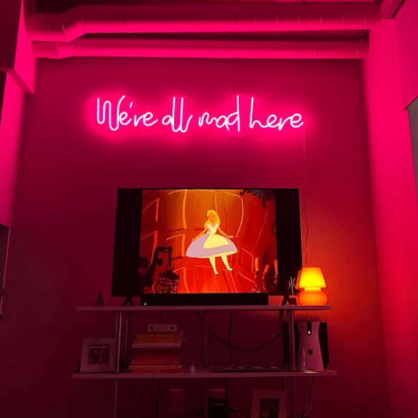 we're all mad here neon sign in deep pink installed on wall above tv. Tv is playing the film "Alice in Wonderland"