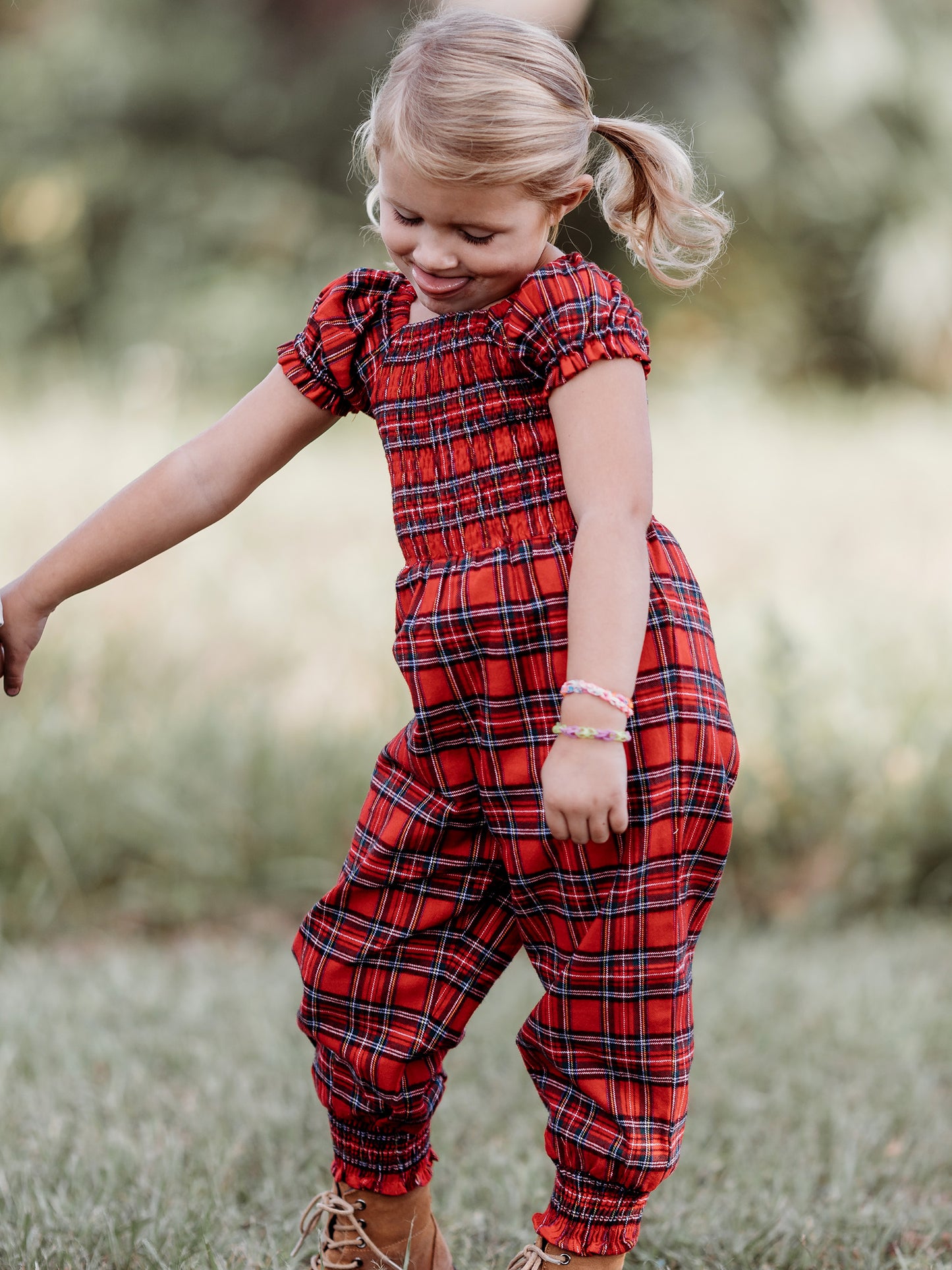 This image of a girl features the product Smocked Romper - Holiday Red Plaid. It comes in a red plaid pattern with accents of white, green, blue, and yellow.