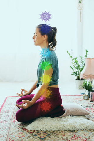 A woman meditates in a kneeling position with the chakra points highlighted with their respective Sanskrit symbols in a rainbow of colours