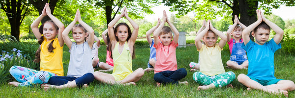 A row of children sat cross legged with their hands in prayer pose doing yoga outside on the grass