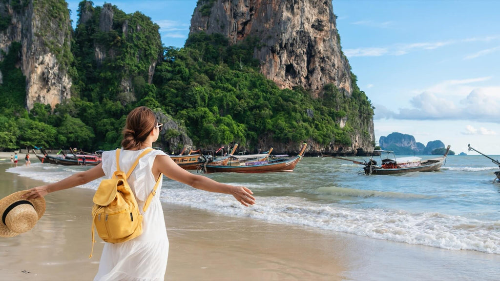 A woman in white dress with yellow backpack spreads her arms wide overlooking a beach with mountains in Thailand