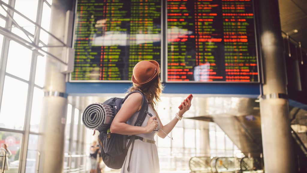 A woman wearing a cream dress and orange backpack holds a pallet and looks at a flight departures board full of cancelled flights