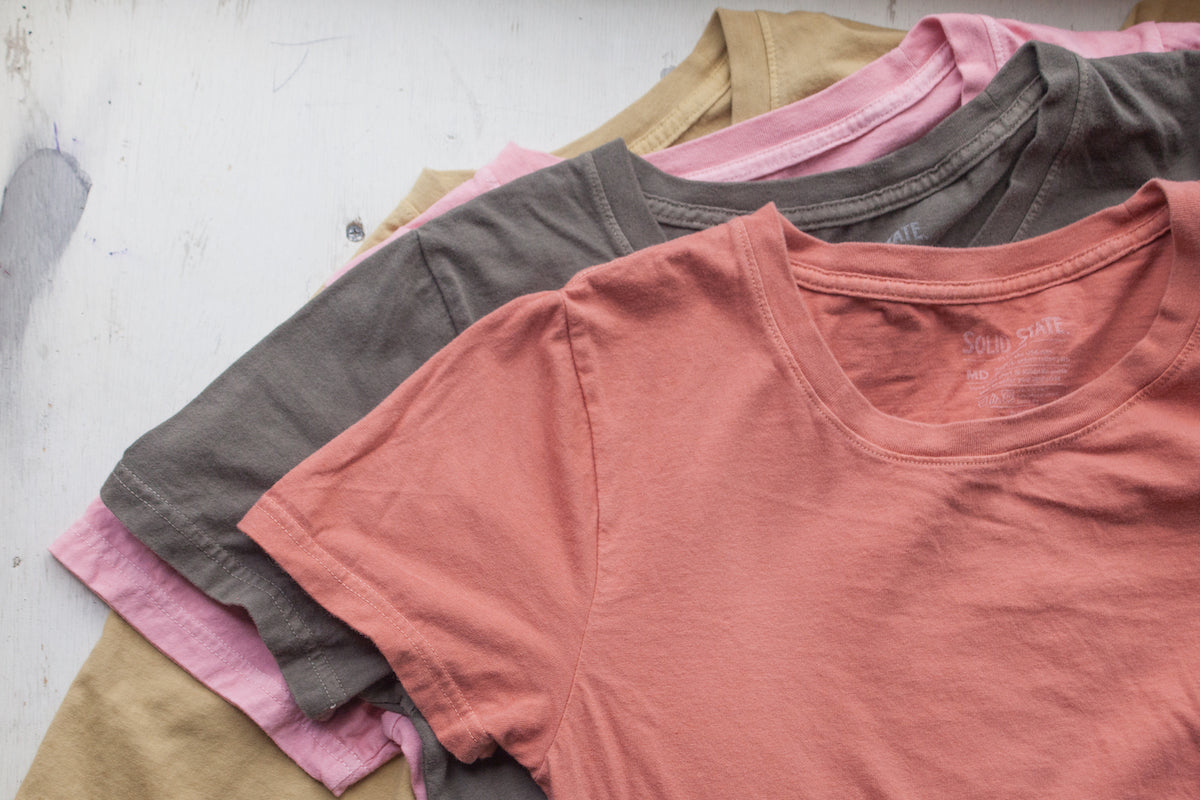 Four t-shirts stacked dyed with natural dyes in orange, green, pink, and yellow