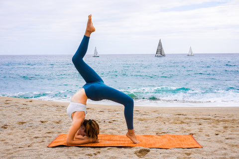 Yoga girl doing a backbend pose on an orange organic cotton yoga mat with the ocean in the background.