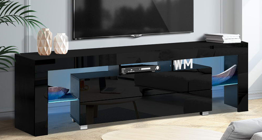 An Artiss Black Gloss RGB LED entertainment unit, holding a DVD player, two decorative bowls and the letters WM. On top is a stack of books and two vases. Above the unit is a wall mounted plasma TV. There is a houseplant dangling over the unit from the left corner.