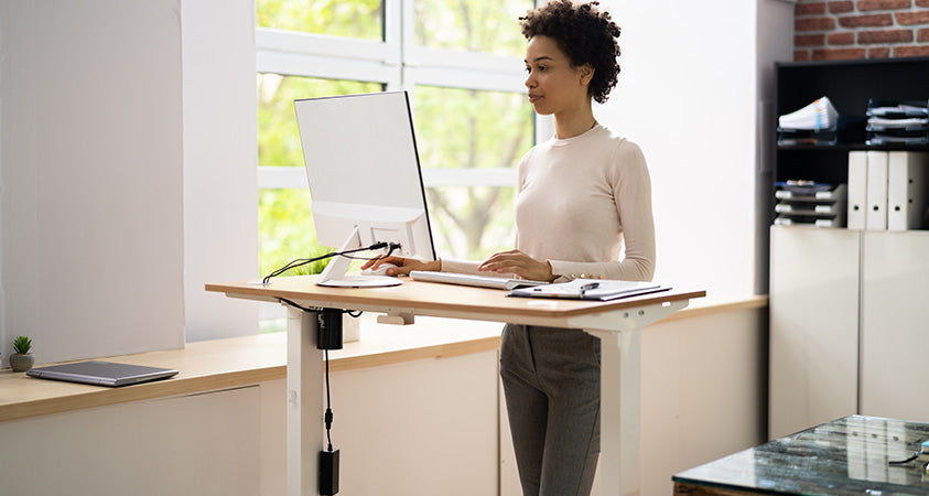 A woman standing in front of an electric standing desk working on her white computer. The standing desk has a wood top and white metal legs.