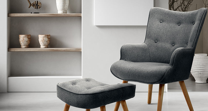 A grey chair and ottoman set against a white wall, near some light wood grain shelving and a white vase. 