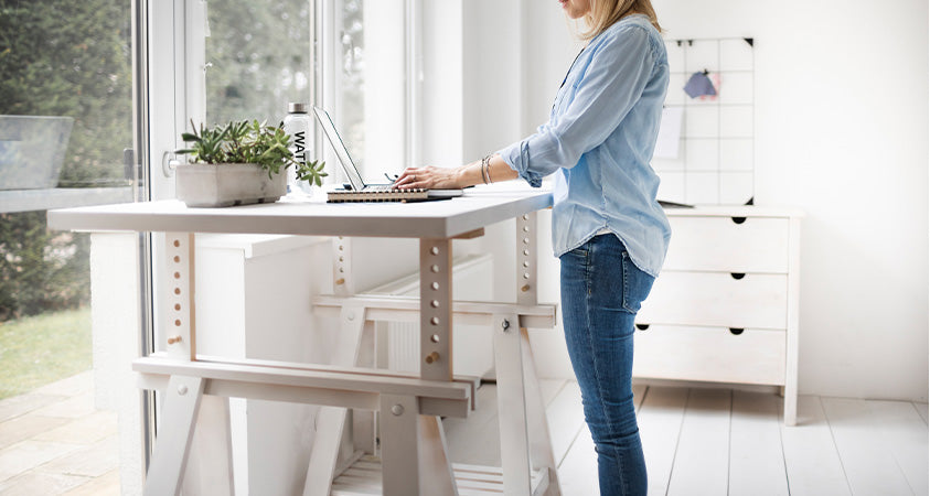 A woman is standing in front of a white manual standing desk, working on a laptop. The space is white with minimal decor, except for a pot plant and water bottle