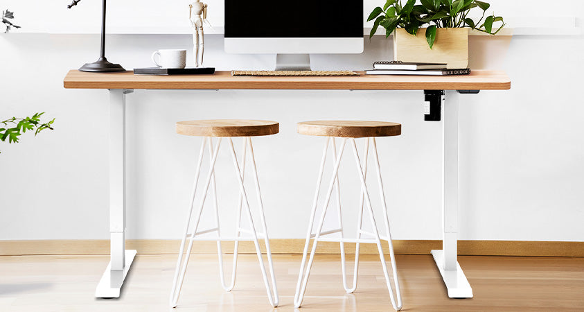 A pneumatic standing desk, with white metal legs and a wood top. The desk is covered with home office supplies including a laptop, lamp, books, pot plant and artists mannequin. There are two stools in front with wood tops and white metal legs. The desk is centered on a white wall with a wood grain floor.