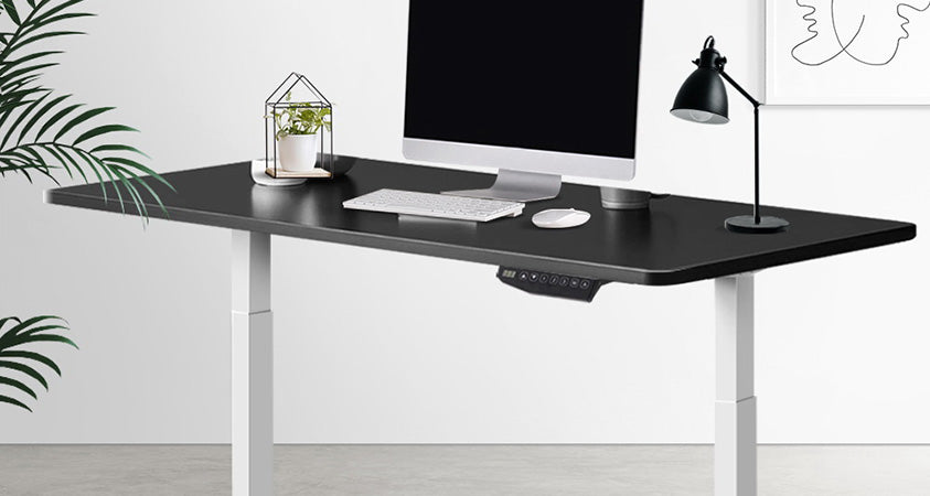 An electric standing desk with white legs and a black top. The desk holds a computer monitor, pot plant, lamp, keyborad and mouse. It is in a minmal office space with a white wall, grey flooring, an abstract art piece and a houseplant.