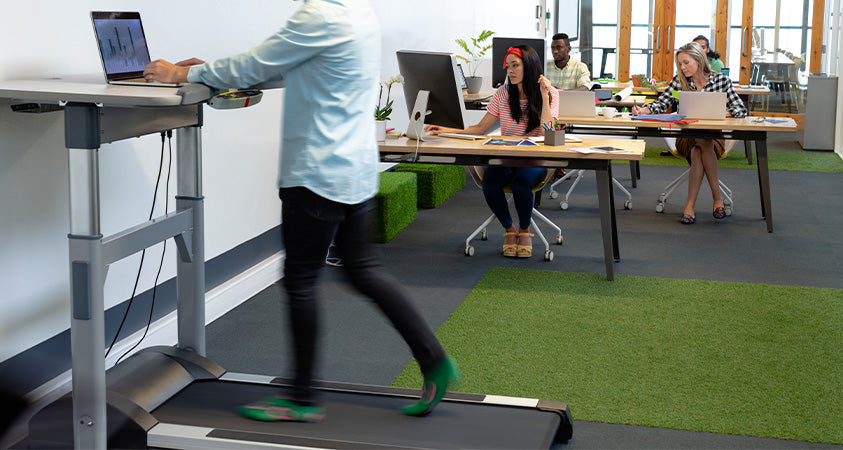 A man is standing at a treadmill desk, walking while working on a laptop. In the background is a busy office with several other workers at normal desks, working behind computers. The floor is mostly grey carpet, broken by two faux-grass rugs and some faux-grass seats.