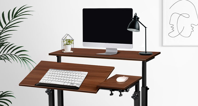 A standing desk with adjustable keyboard and mouse holders. The desk has a dark wood top and black legs. The keyboard is tilted slightly towards the viewer, while the mouse is flat. The desk also features a computer monitor, pot plant and lamp. The desk is in a white room with a single abstract painting and a houseplant.