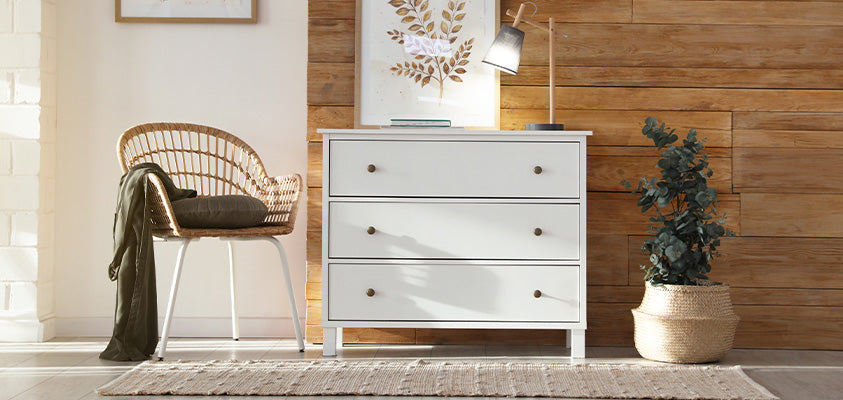 A simple white 3-drawer chest of drawers set against a half wood, half beige wall. On the left is a woven chair with a cushion and blanket, on the left is a pot plant in a woven basket. The drawers are holding some natural art, notebooks and a lamp.