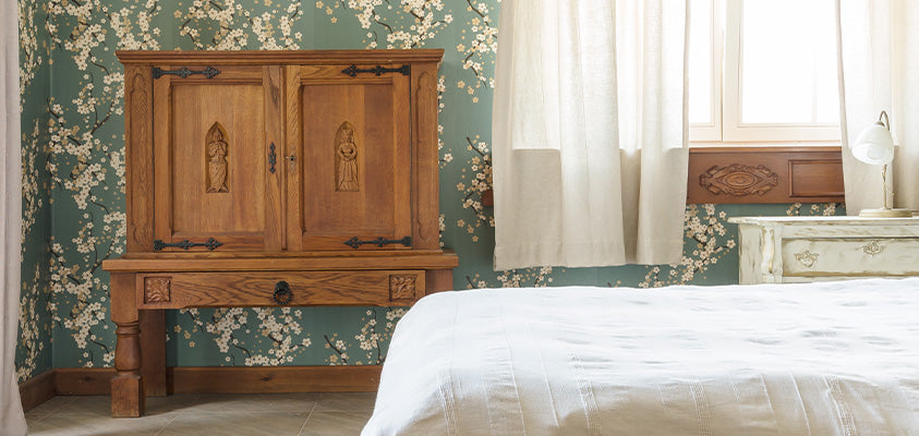 A bedroom covered in floral wallpaper. In the corner is a wooden cabinet with a single drawer. It has several unique carvings. To the left is a rustic white beside table, an open window with an ornately carved windowsill, and the end of a bed.