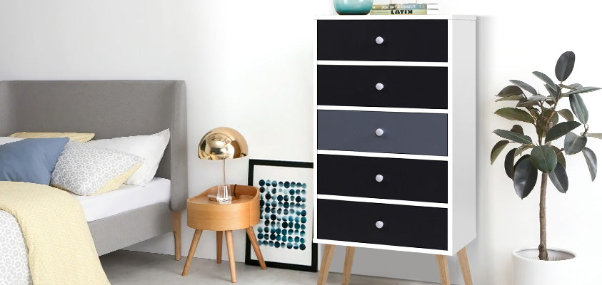 The Artss 5-drawer, 3 tone tallboy with white knobs and light wood grain legs. It is set in a bright bedroom with grey, blue, yellow and white tones.
