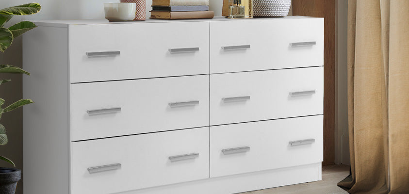 A 6-drawer white chest of drawers with silver handles, set in the corner of a room between a window and a pot plant.