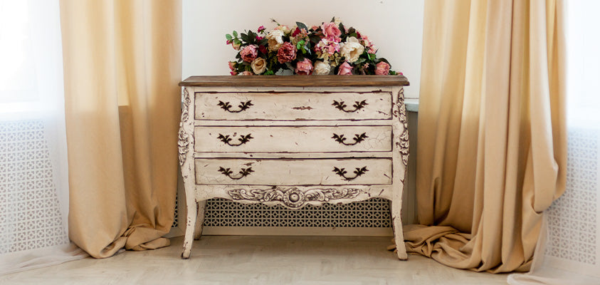 A rustic provincial style chest of drawers. It is white with distressed paint and has a solid wood top and has decorative black metal handles. It is set between two cream coloured curtains.