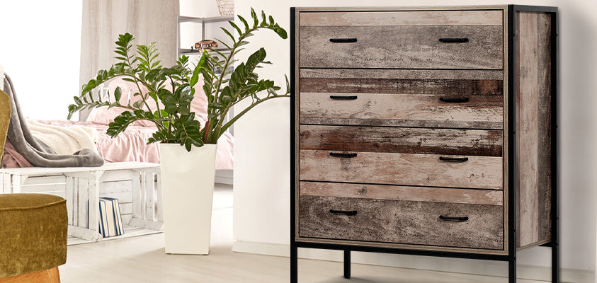 An Artiss 4 drawer Barnsly tallboy in black metal and wood. It is set around the corner from a light bedroom setting, with a large green pot plant directly on the right.
