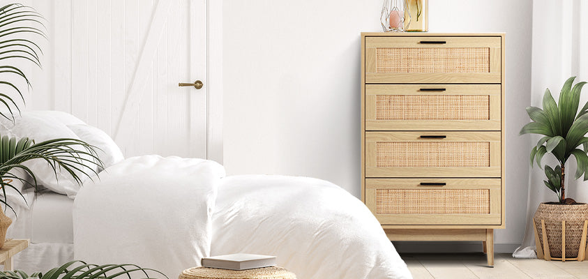 An Artiss 4-drawer ratten chest of drawers. It is set between a white bed and a woven basket with a plant. The chest of drawers has a candle, candle holder and vase set on top. 