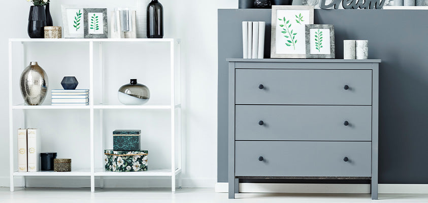 A set of white open metal shelves against a white wall, next to a blue 3-drawer tallboy against a blue wall. Each is holding various household objects such as glass vases, books, decorative boxes and artwork of plants.