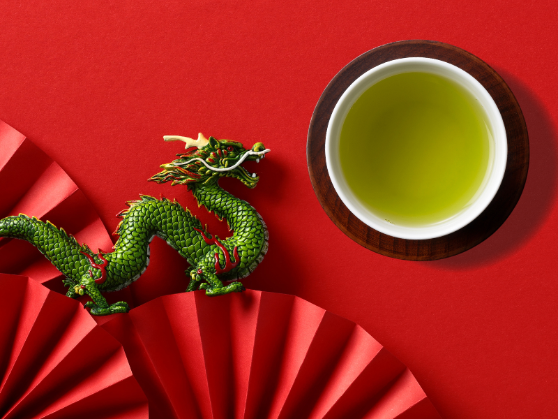 green dragon on red background with red paper fans. nesxt to white teacup with green tea on wood coaster