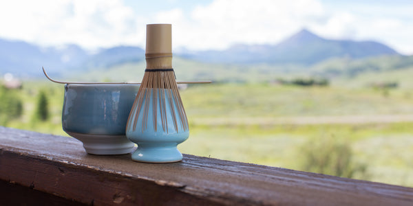 tazón de matcha azul cielo y soporte para batir con batidor en una cornisa de madera con vista a campos verdes y montañas en la distancia