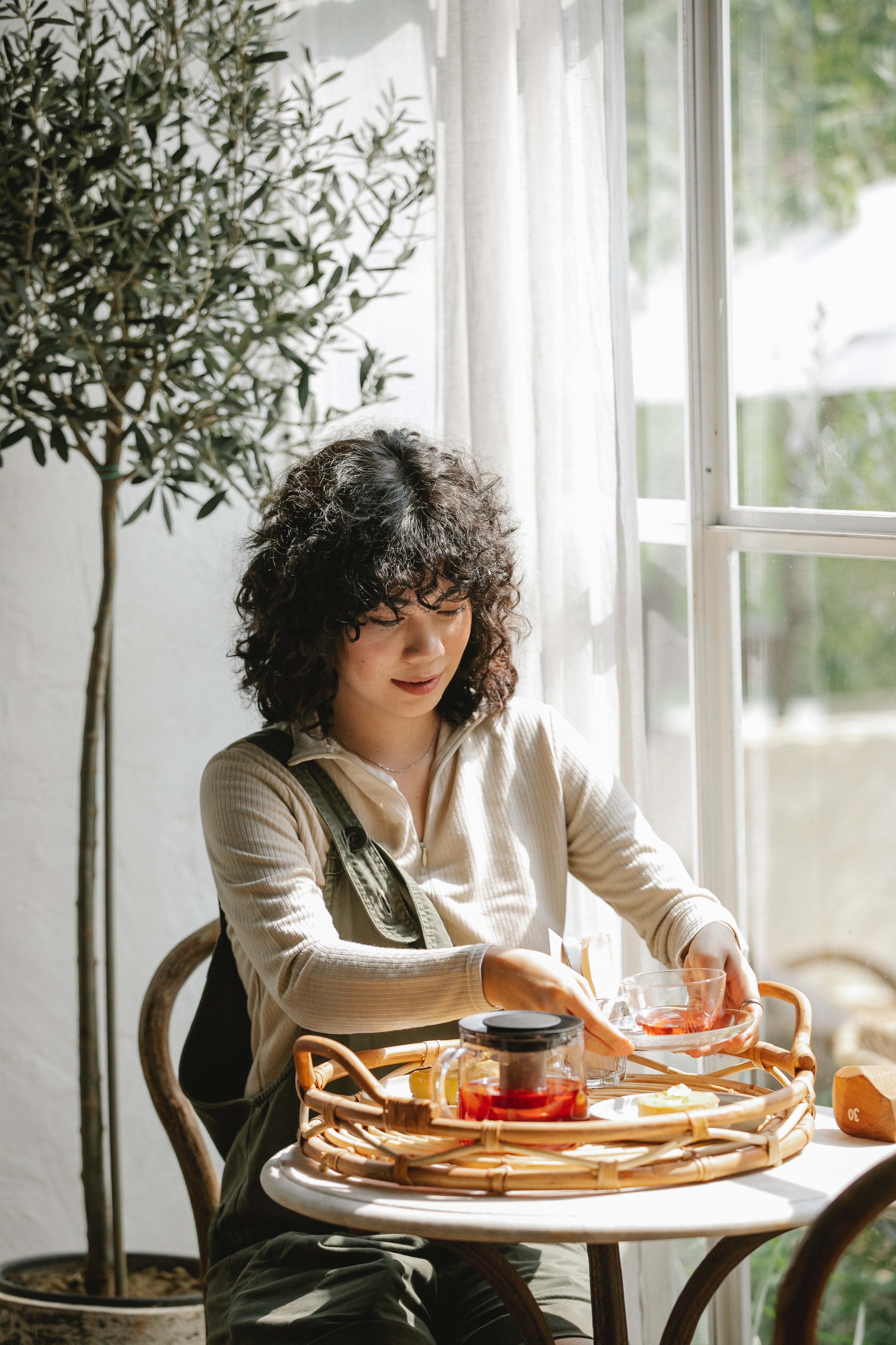 brightly lit cafe interior. Small table next to window with young woman and a tray and teapot with tea