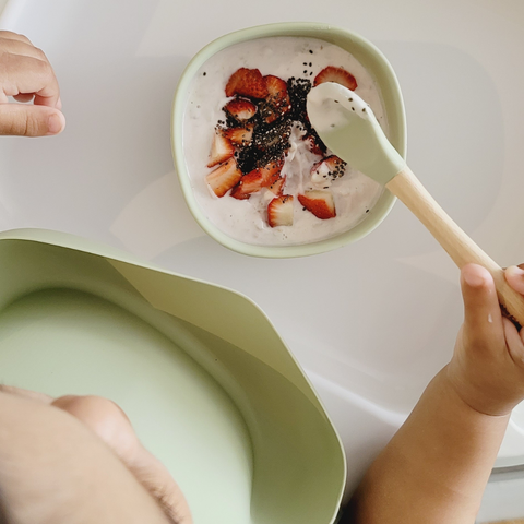 baby eating fruits and yogurt from his silicone bowl while using a silicone spoon and wearing a silicone bib