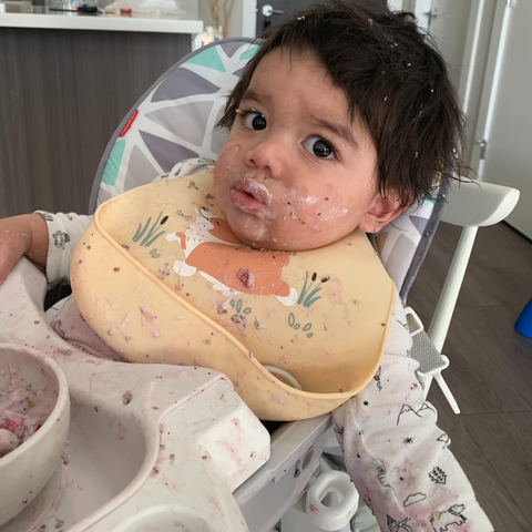 baby wearing a silicone bib while having food smeared all over his face and hair