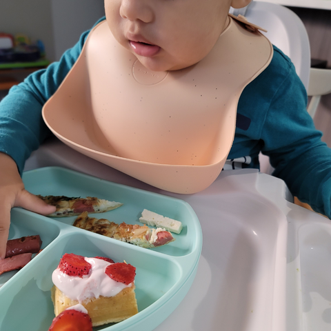 Baby wearing silicone bib while using his hands to eat his meal from a silicone divided plate for kids