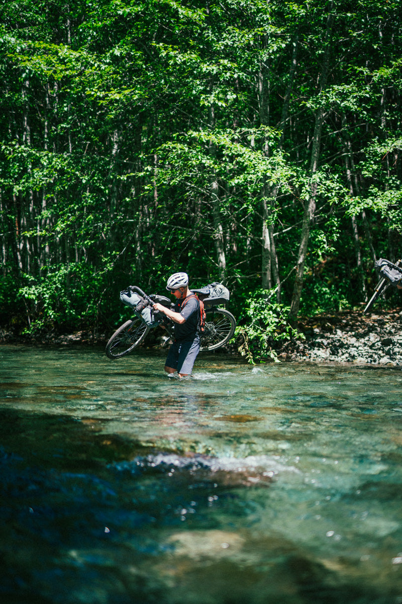 Colin wades through a river with his bike on his shoulder