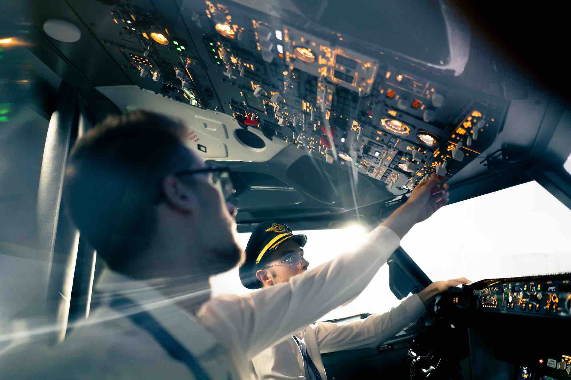 Two pilots in cockpit with the nearest pilot reaching for controls on the overhead boards.