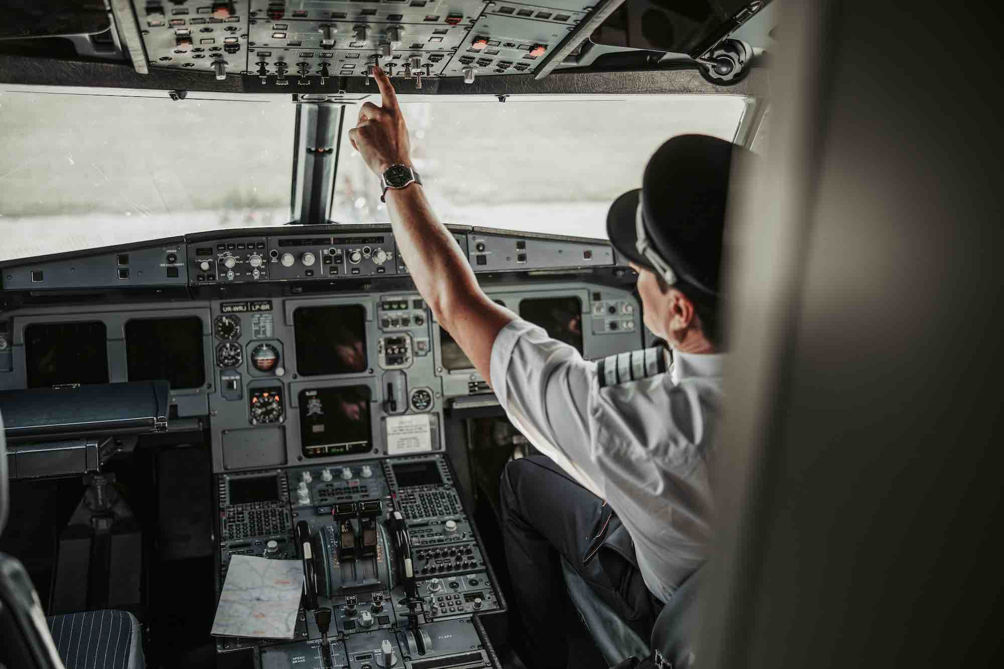 Pilot in cockpit reaching for buttons on overhead boards.